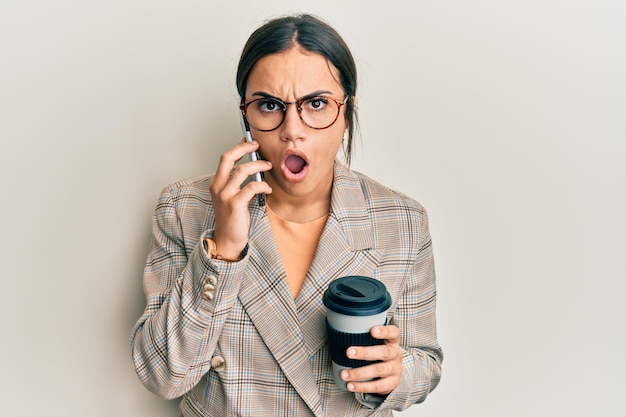 Giovane donna castana che usa lo smartphone e beve una tazza di caffè impaurita e scioccata dalla sorpresa e dall'espressione stupita paura e viso eccitato