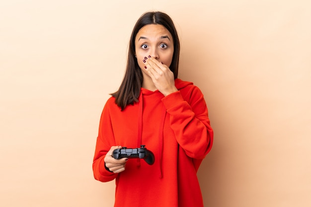Giovane donna castana della corsa mista che gioca con un regolatore del videogioco sopra la parete isolata con espressione facciale di sorpresa