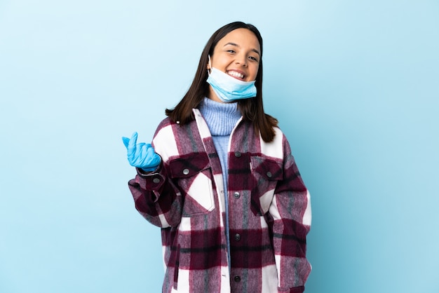 Giovane donna castana della corsa mista che protegge con una maschera e guanti sopra la parete blu che fa gesto dei soldi