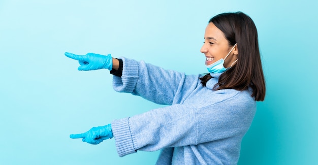 Giovane donna castana della corsa mista che protegge con una maschera e guanti sopra la parete blu che indica dito il lato e che presenta un prodotto