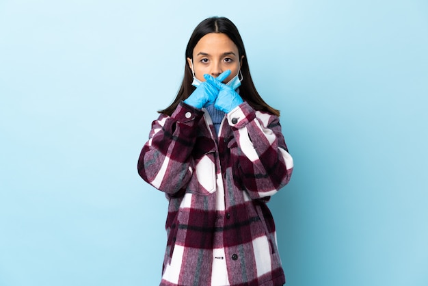 Giovane donna castana della corsa mista che protegge con una maschera e guanti sopra la parete blu che mostra un segno di gesto di silenzio