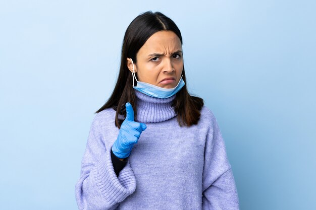 Giovane donna castana della corsa mista che protegge con una maschera e guanti sopra la parete blu frustrata e indicante