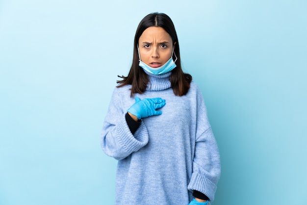 Giovane donna castana della corsa mista che protegge dal coronavirus con una maschera e guanti sopra la parete blu isolata che indica a se stessi