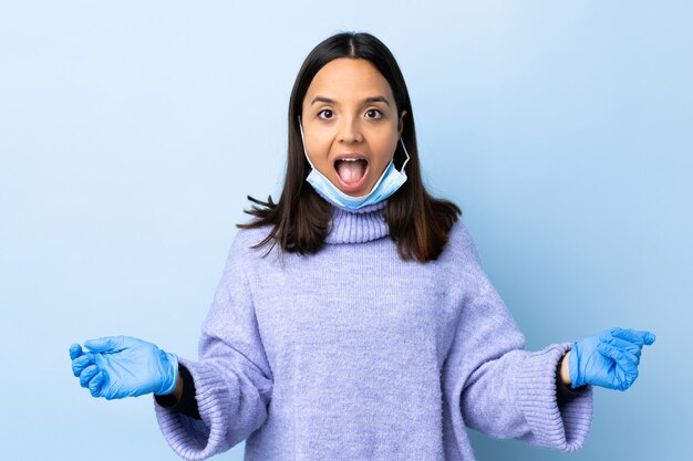 Giovane donna castana della corsa mista che protegge dal coronavirus con una maschera e guanti sopra la parete blu isolata con espressione facciale di sorpresa