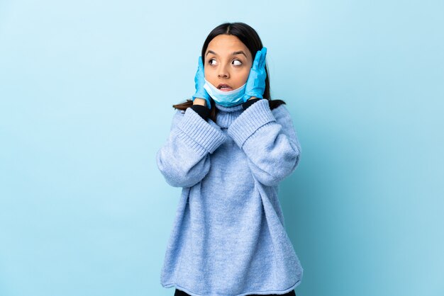 Giovane donna castana della corsa mista che protegge dal coronavirus con una maschera e guanti sopra la parete blu isolata frustrata e che copre le orecchie