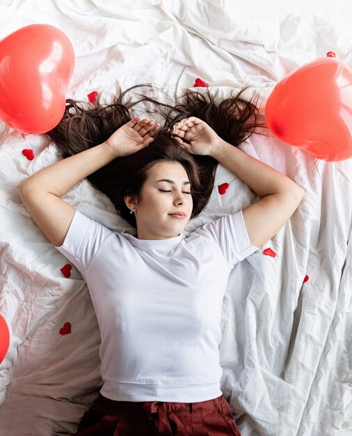 Giovane donna castana felice che risiede nel letto con palloncini e decorazioni a forma di cuore rosso