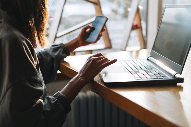 Giovane donna castana in abbigliamento casual che lavora con il computer portatile utilizzando il telefono cellulare al caffè