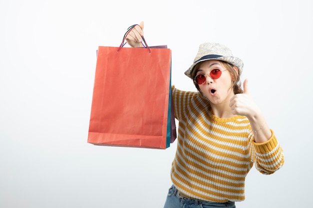 Giovane donna castana in occhiali da sole che tiene le borse della spesa e mostra il pollice in su. Foto di alta qualità
