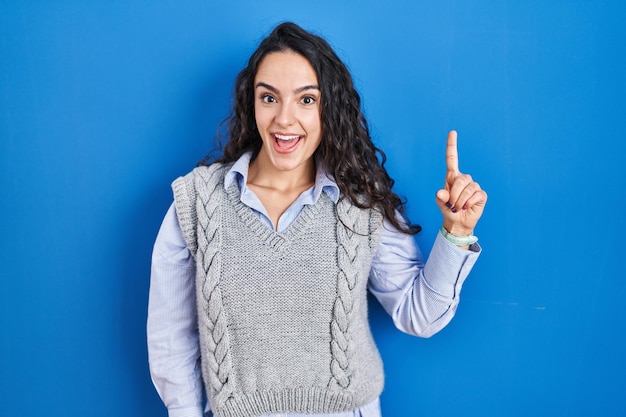 Giovane donna castana in piedi su sfondo blu che punta il dito verso l'alto con un'idea di successo uscita e felice numero uno