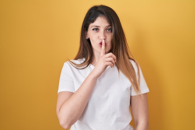 Giovane donna castana in piedi su sfondo giallo che chiede di stare zitta con il dito sulle labbra silenzio e concetto segreto