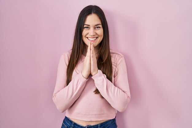 Giovane donna castana in piedi su sfondo rosa che prega con le mani insieme chiedendo perdono sorridendo fiducioso