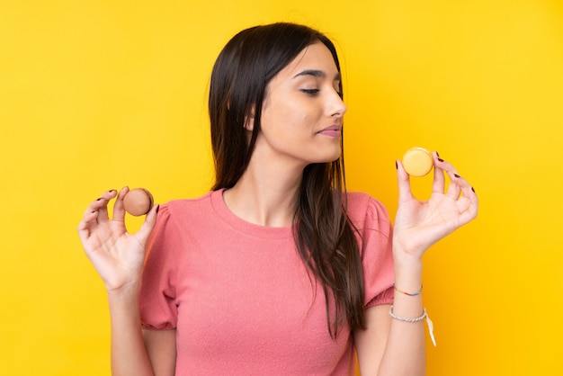 Giovane donna castana sopra la parete gialla isolata che tiene i macarons francesi variopinti e che lo guarda