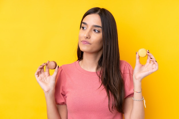 Giovane donna castana sopra la parete gialla isolata che tiene macarons e pensiero francesi variopinti