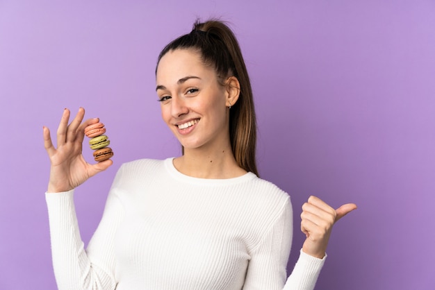 Giovane donna castana sopra la parete porpora isolata che tiene i macarons francesi variopinti e che indica lato