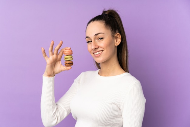 Giovane donna castana sopra la parete porpora isolata che tiene i macarons francesi variopinti e che sorride molto