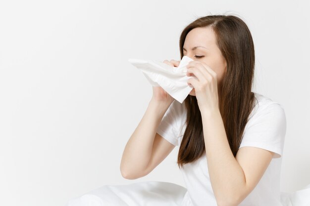 Giovane donna castana stanca di malattia in pigiama che si siede sul letto isolato sul wall . bianco