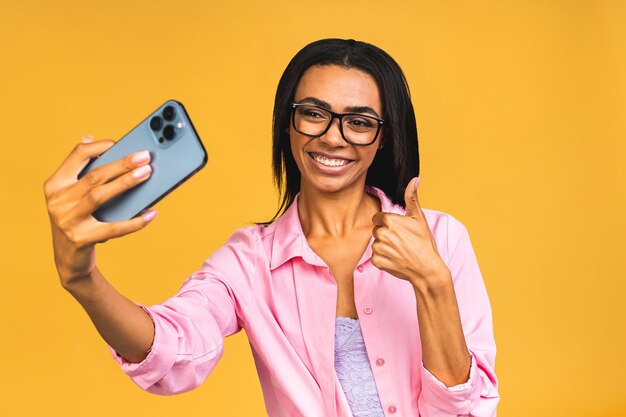 Giovane donna casual nera afroamericana divertente che fa una smorfia e fa selfie sullo smartphone isolato su sfondo giallo