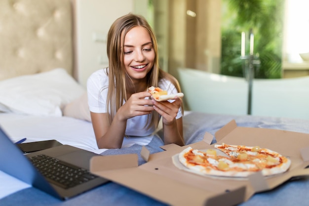 Giovane donna casual sdraiata su un letto a letto a casa mentre mangia pizza e usa il laptop