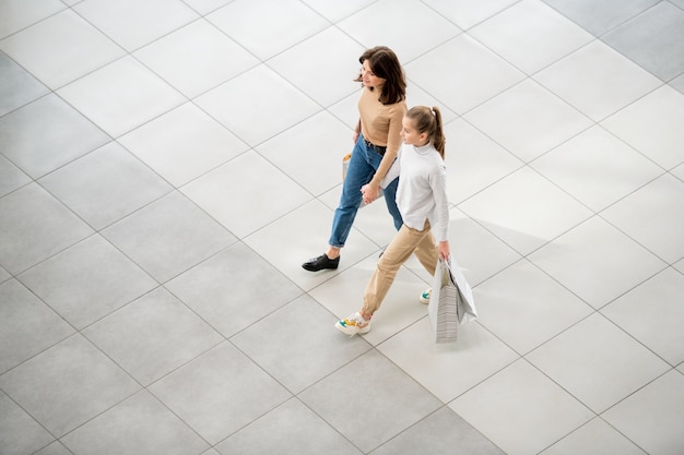 Giovane donna casuale e sua figlia con i sacchetti di carta che si muovono lungo il centro commerciale contemporaneo mentre fanno shopping