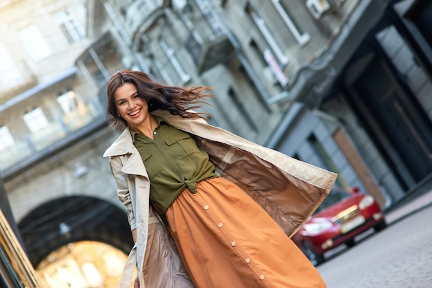 Giovane donna caucasica alla moda eccitata che indossa un cappotto grigio che guarda l'obbiettivo e sorride mentre sta in piedi