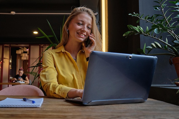Giovane donna caucasica attraente ben vestita che si siede al tavolo con il computer portatile che chiama dallo smartphone
