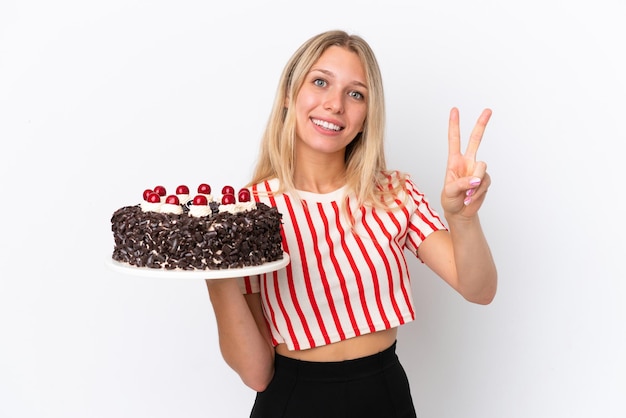 Giovane donna caucasica azienda torta di compleanno isolata su sfondo bianco sorridente e mostrando il segno di vittoria