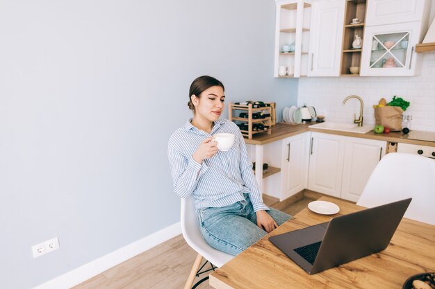 Giovane donna caucasica bere caffè e utilizzando il computer portatile mentre è seduto su una sedia in cucina moderna.