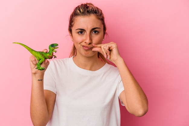 Giovane donna caucasica bionda che tiene una miniatura di dinosauro isolata sulla parete rosa con le dita sulle labbra mantenendo un segreto