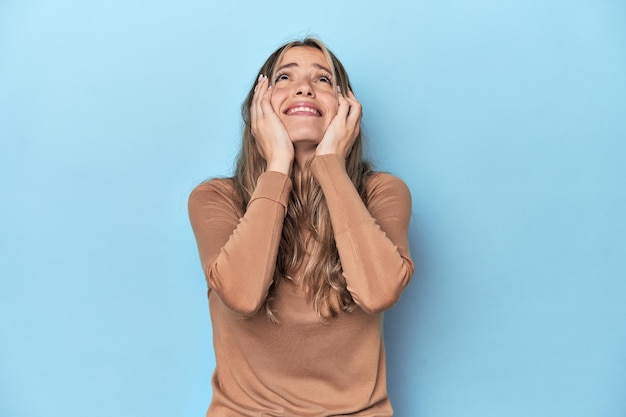 Giovane donna caucasica bionda in studio blu che piange insoddisfatta di qualcosa di agonia