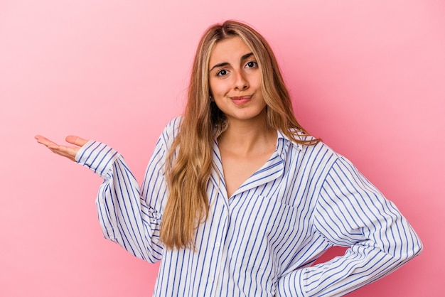 Giovane donna caucasica bionda isolata sulla parete rosa dubitando e alzando le spalle nel gesto interrogativo.