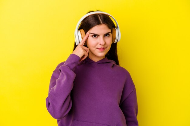 Giovane donna caucasica che ascolta musica con le cuffie isolate su sfondo rosa che punta il tempio con il dito, pensando, concentrato su un compito.