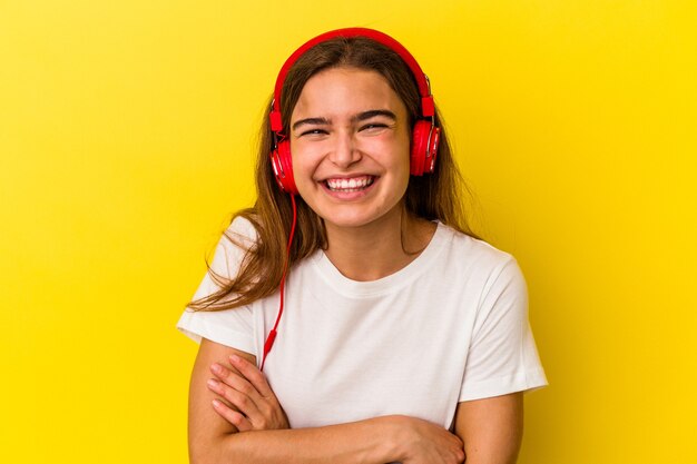 Giovane donna caucasica che ascolta musica isolata su sfondo giallo ridendo e divertendosi.