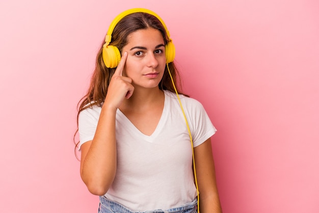 Giovane donna caucasica che ascolta musica isolata su sfondo rosa che punta il tempio con il dito, pensando, concentrata su un compito.