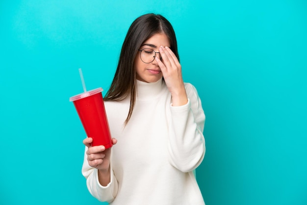 Giovane donna caucasica che beve soda isolata su sfondo blu con espressione stanca e malata