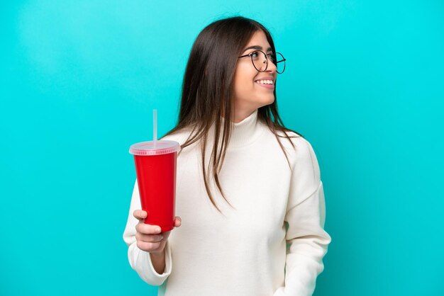 Giovane donna caucasica che beve soda isolata su sfondo blu guardando di lato e sorridente