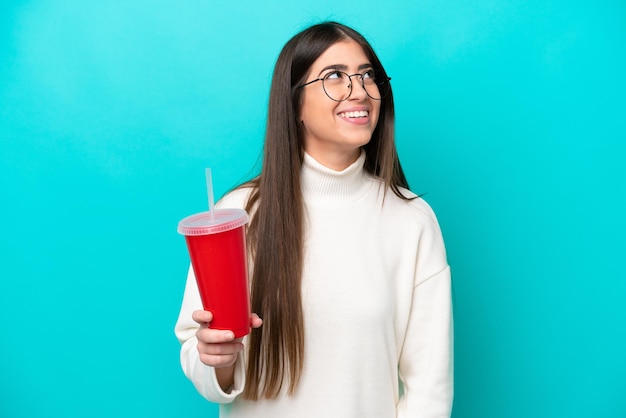 Giovane donna caucasica che beve soda isolata su sfondo blu pensando un'idea mentre cerca