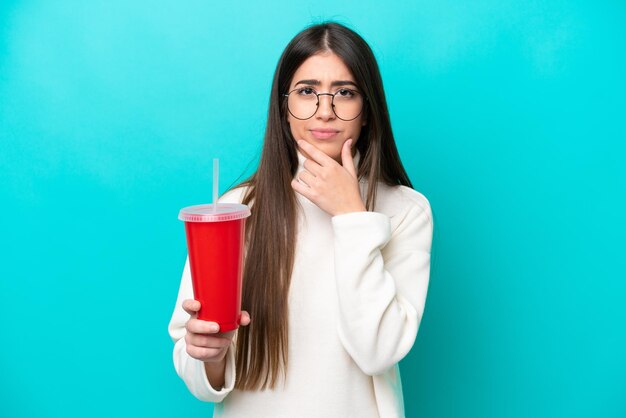 Giovane donna caucasica che beve soda isolata su sfondo blu pensando