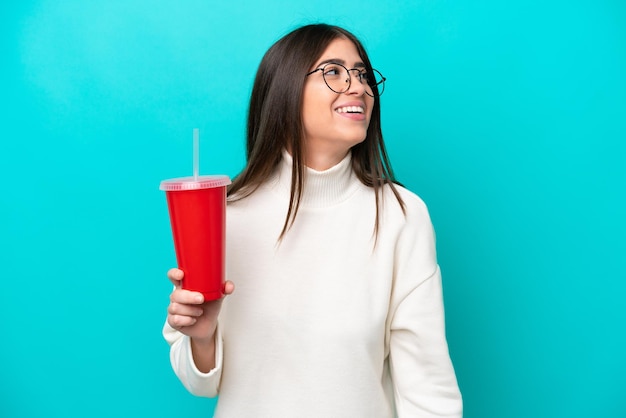 Giovane donna caucasica che beve soda isolata su sfondo blu ridendo