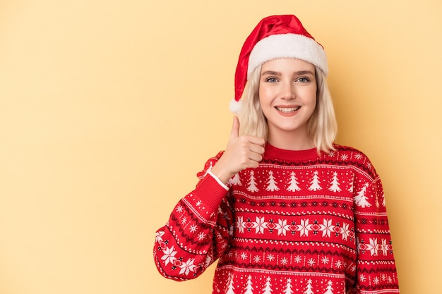 Giovane donna caucasica che celebra il Natale isolato su sfondo giallo sorridente e alzando il pollice