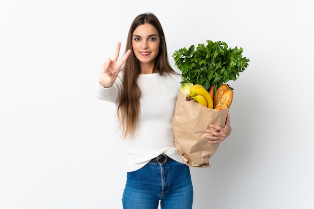 Giovane donna caucasica che compra del cibo isolato sulla parete bianca che sorride e che mostra il segno di vittoria