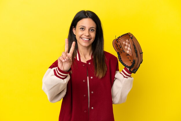 Giovane donna caucasica che gioca a baseball isolata su fondo giallo che sorride e che mostra il segno di vittoria