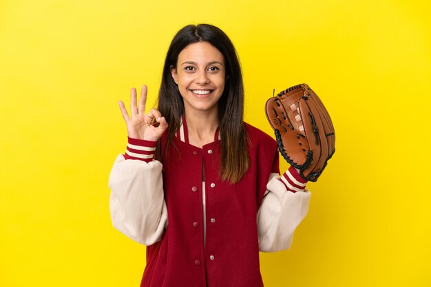 Giovane donna caucasica che gioca a baseball isolata su sfondo giallo che mostra segno ok con le dita