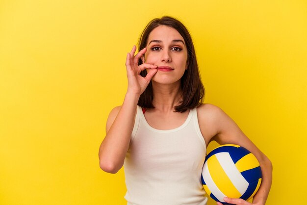 Giovane donna caucasica che gioca a pallavolo isolata su sfondo giallo con le dita sulle labbra mantenendo un segreto