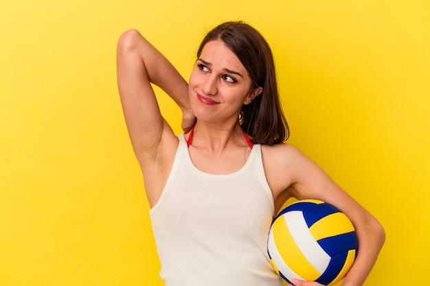 Giovane donna caucasica che gioca a pallavolo isolata su sfondo giallo toccando la parte posteriore della testa, pensando e facendo una scelta.