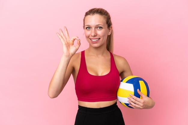 Giovane donna caucasica che gioca a pallavolo isolata su sfondo rosa che mostra il segno giusto con le dita