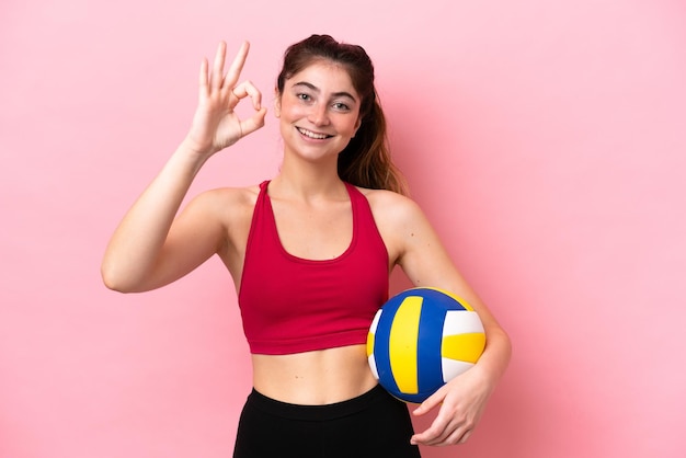 Giovane donna caucasica che gioca a pallavolo isolata su sfondo rosa che mostra il segno giusto con le dita