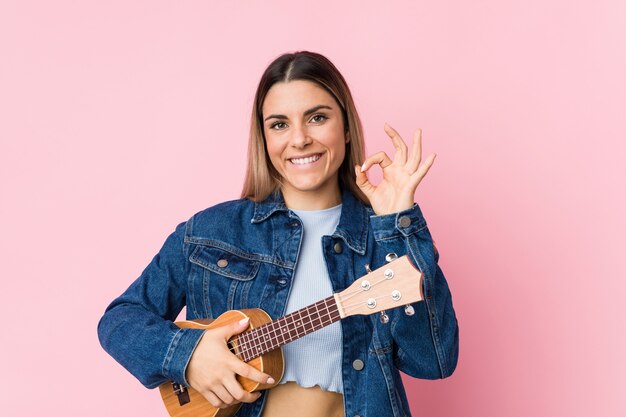 Giovane donna caucasica che gioca ukelele allegro e fiducioso mostrando gesto ok.