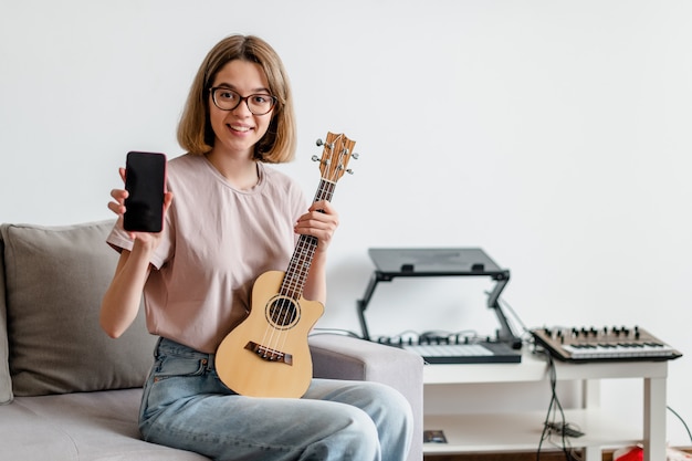 Giovane donna caucasica che gioca ukulele utilizzando l'app mobile a casa studio