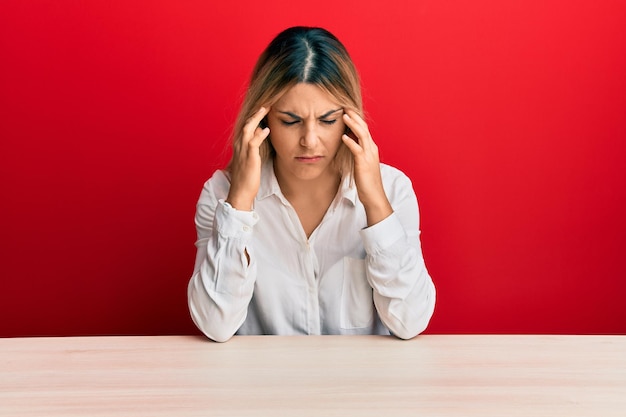 Giovane donna caucasica che indossa abiti casual seduta sul tavolo con la mano sulla testa per il dolore alla testa perché lo stress soffre di emicrania