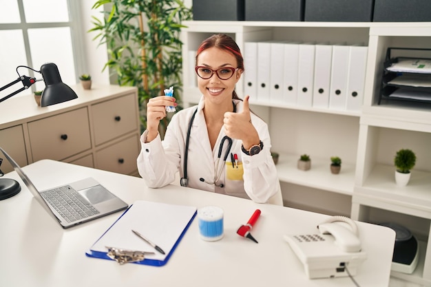 Giovane donna caucasica che indossa l'uniforme del medico che tiene i cotton fioc sorridendo felice e positivo, pollice in su facendo eccellente e segno di approvazione